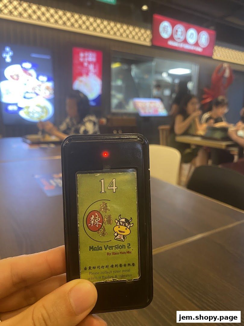 A hand holds a restaurant pager with the shop name 'Mala Version 2' visible.