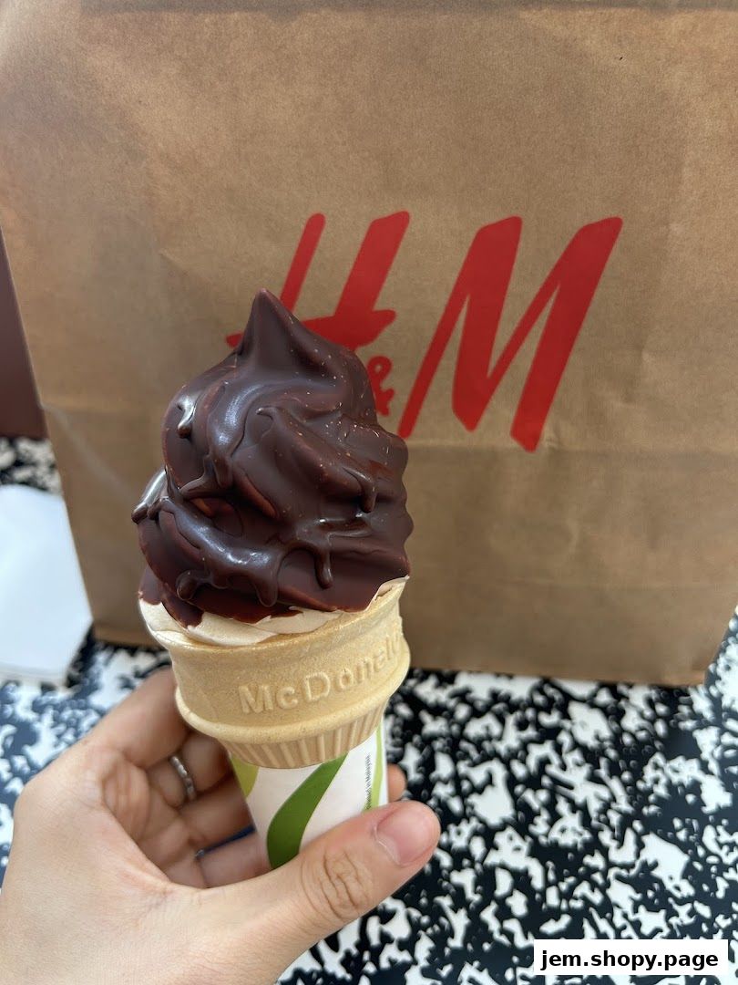 A hand holds a chocolate-dipped ice cream cone in front of a brown shopping bag.