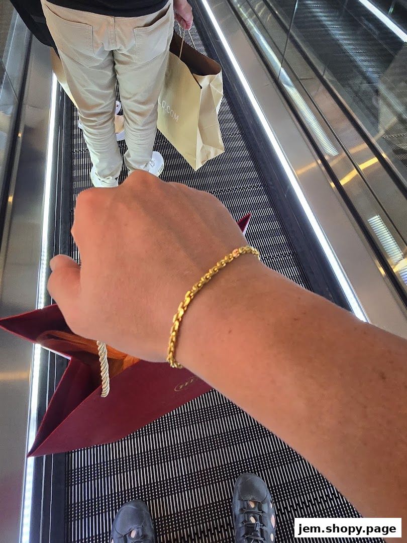 A person wearing a gold bracelet and holding a shopping bag on an escalator.