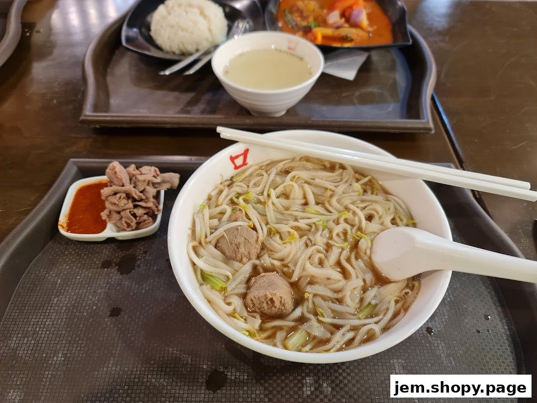 A bowl of beef noodles with side dishes and a bowl of rice on a tray.