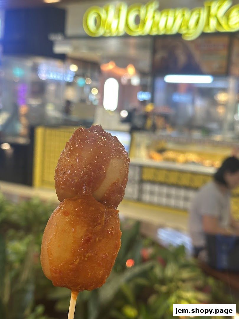 A close-up of a skewer with two savory snacks, with the Old Chang Kee logo blurred in the background.