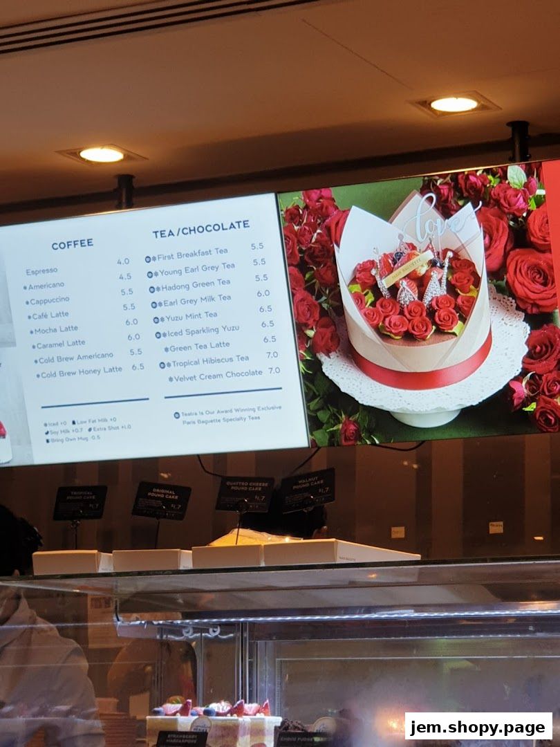 A digital menu board displaying coffee and tea options with prices, alongside a cake display.