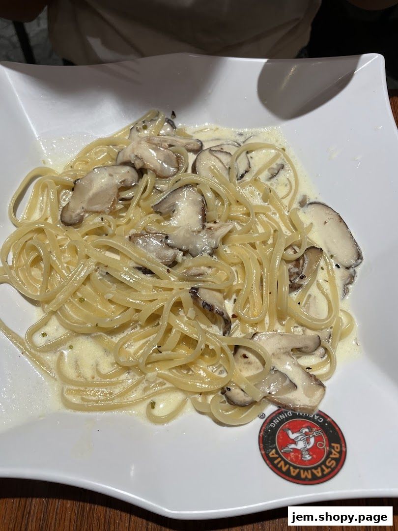 A plate of creamy pasta with mushrooms, served in a white square dish with the PastaMania logo.