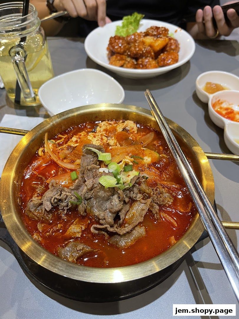 A close-up of a steaming Korean stew with beef, noodles, and kimchi, served with side dishes.