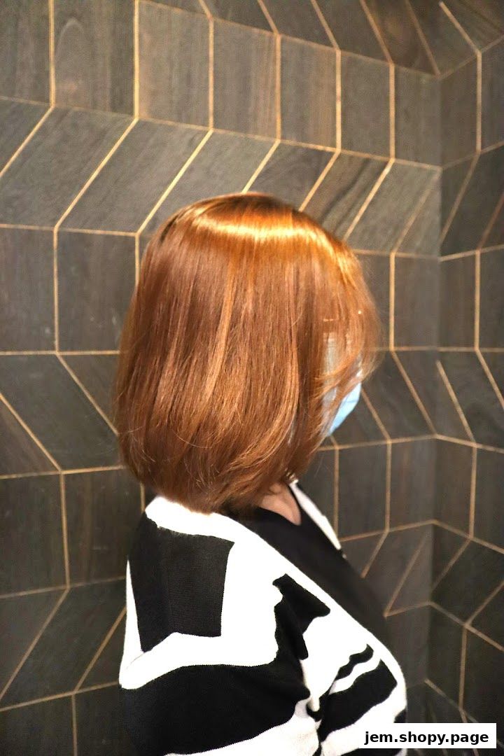 A person with a stylish auburn bob haircut and a face mask stands against a geometric tiled wall.
