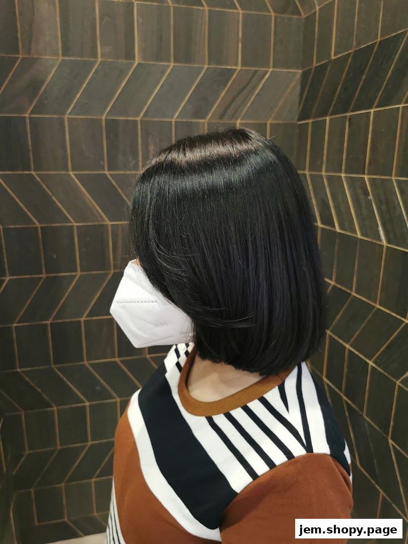A person with a sleek bob haircut and a mask stands against a textured wall.