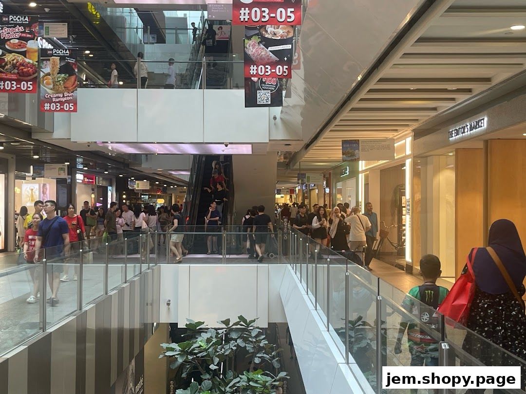 A busy shopping mall interior with escalators, shops, and many shoppers.