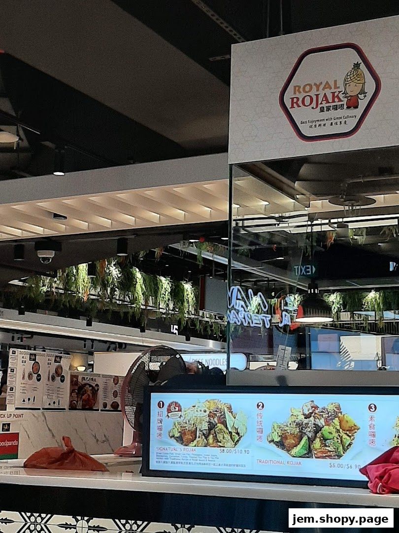 A food stall counter with a menu display showing Rojak dishes and a pick-up point sign.