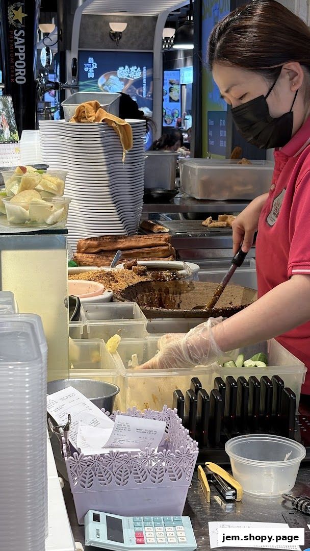 A food stall worker prepares food behind a counter with ingredients and stacks of plates.