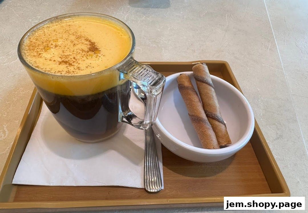 A clear glass mug filled with dark coffee topped with frothy, golden foam and a sprinkle of cinnamon, served with two chocolate wafer rolls.