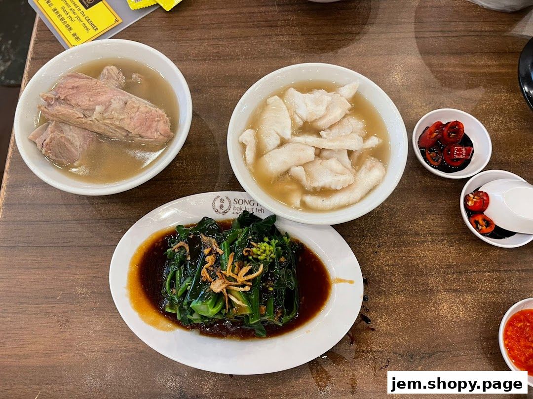 A table setting with bowls of bak kut teh, fish slices, and stir-fried vegetables.