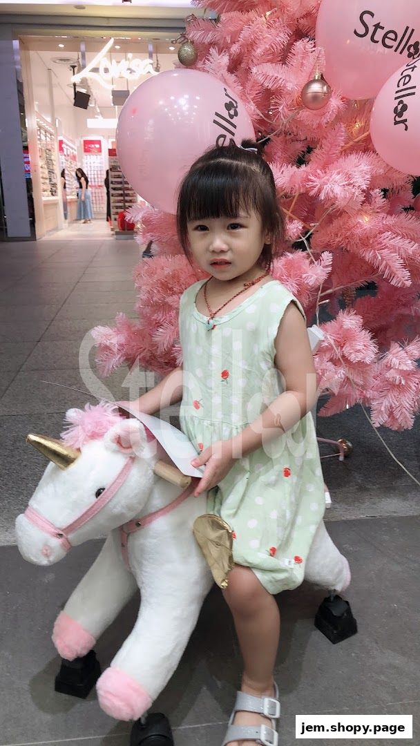 A young girl sits on a unicorn toy in front of a pink Christmas tree with Stellar balloons.