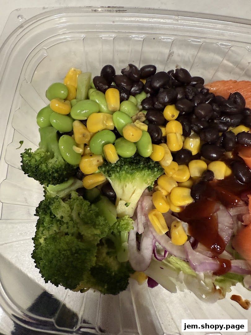 A clear plastic container filled with a fresh salad featuring broccoli, edamame, corn, black beans, and red onion.