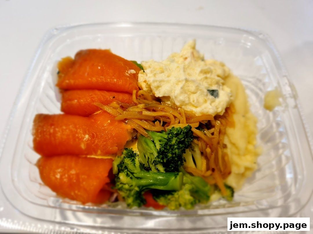A clear plastic container filled with a meal including smoked salmon, mashed potatoes, broccoli, and noodles.