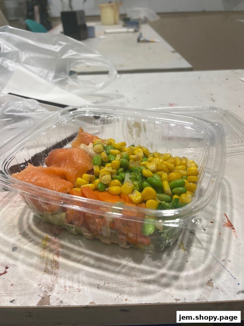 A clear plastic container filled with a colorful salad featuring salmon, corn, edamame, and broccoli.
