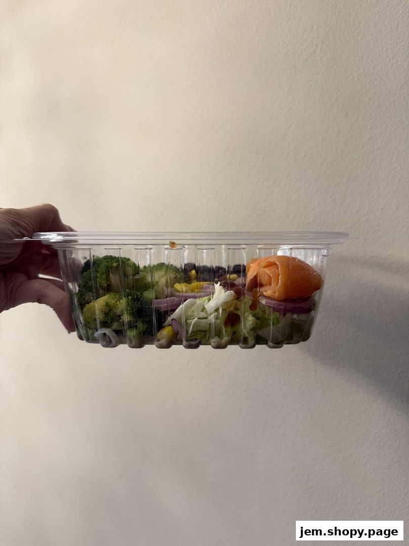 A hand holds a clear plastic container filled with a fresh salad featuring salmon, broccoli, and red onion.