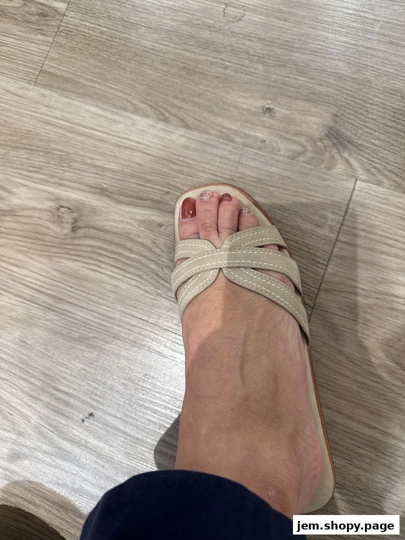 A close-up of a foot wearing a beige sandal with a light wood floor background.