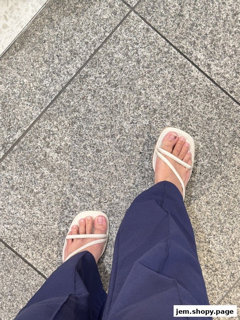 A person wearing white strappy sandals and navy blue pants on a tiled floor.