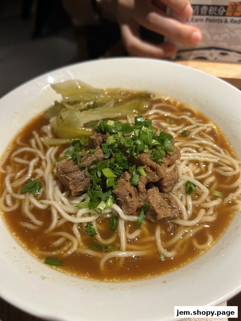 A close-up of a steaming bowl of beef noodle soup with pickled vegetables and scallions.