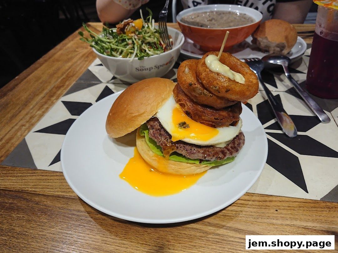 A gourmet burger with onion rings and a fried egg, served with soup and salad.