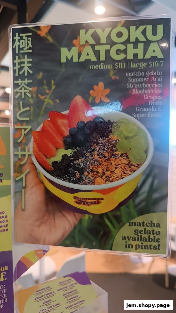 A hand holds a bowl of matcha açaí with fruits and granola, with a menu board behind it.