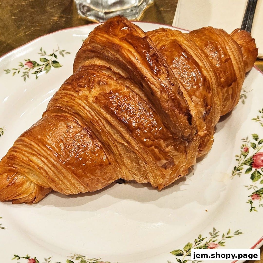 A golden-brown, flaky croissant sits on a decorative plate with floral accents.