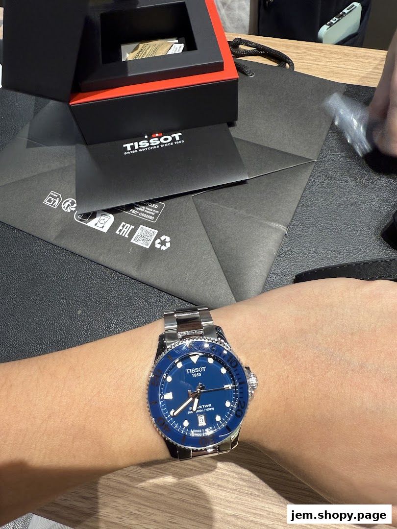A Tissot watch with a blue dial is worn on a wrist, with packaging in the background.