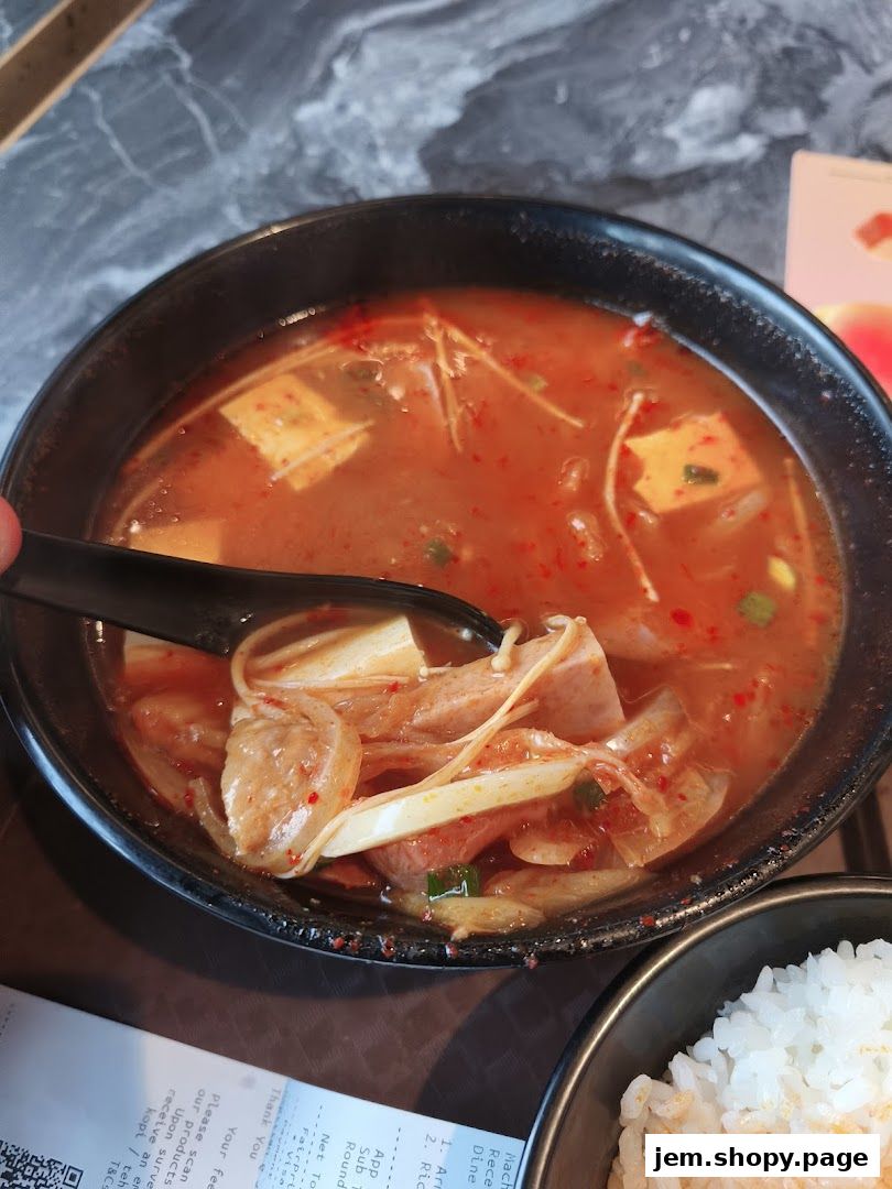 A close-up of a steaming bowl of Korean stew with tofu, mushrooms, and meat, served with rice.