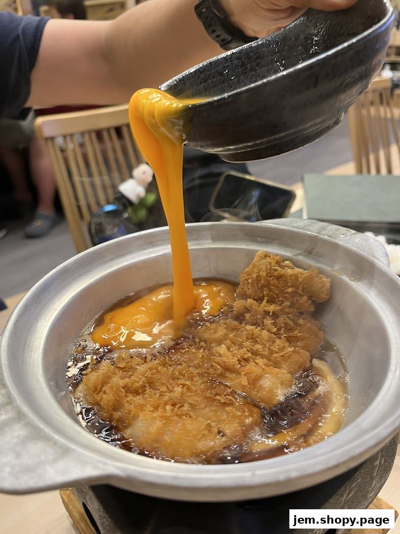 A person pours a rich, golden egg yolk over a steaming bowl of tonkatsu.