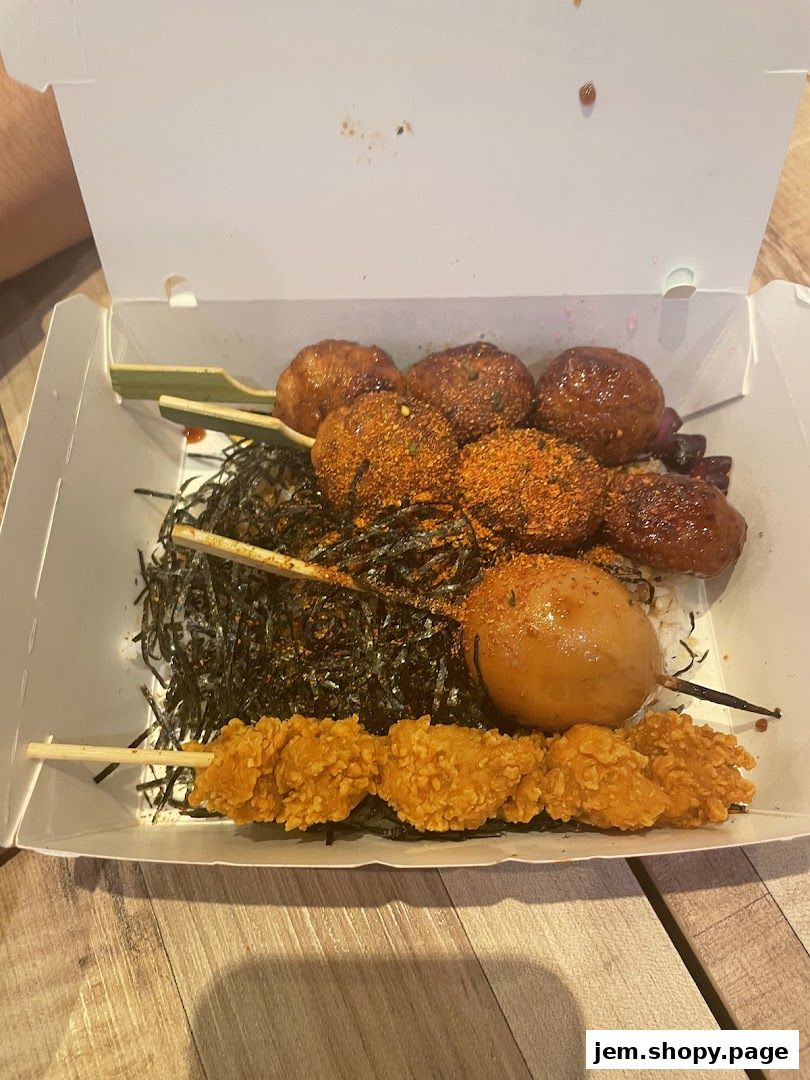 A take-out box filled with rice, seaweed, skewers of meatballs, and fried chicken.