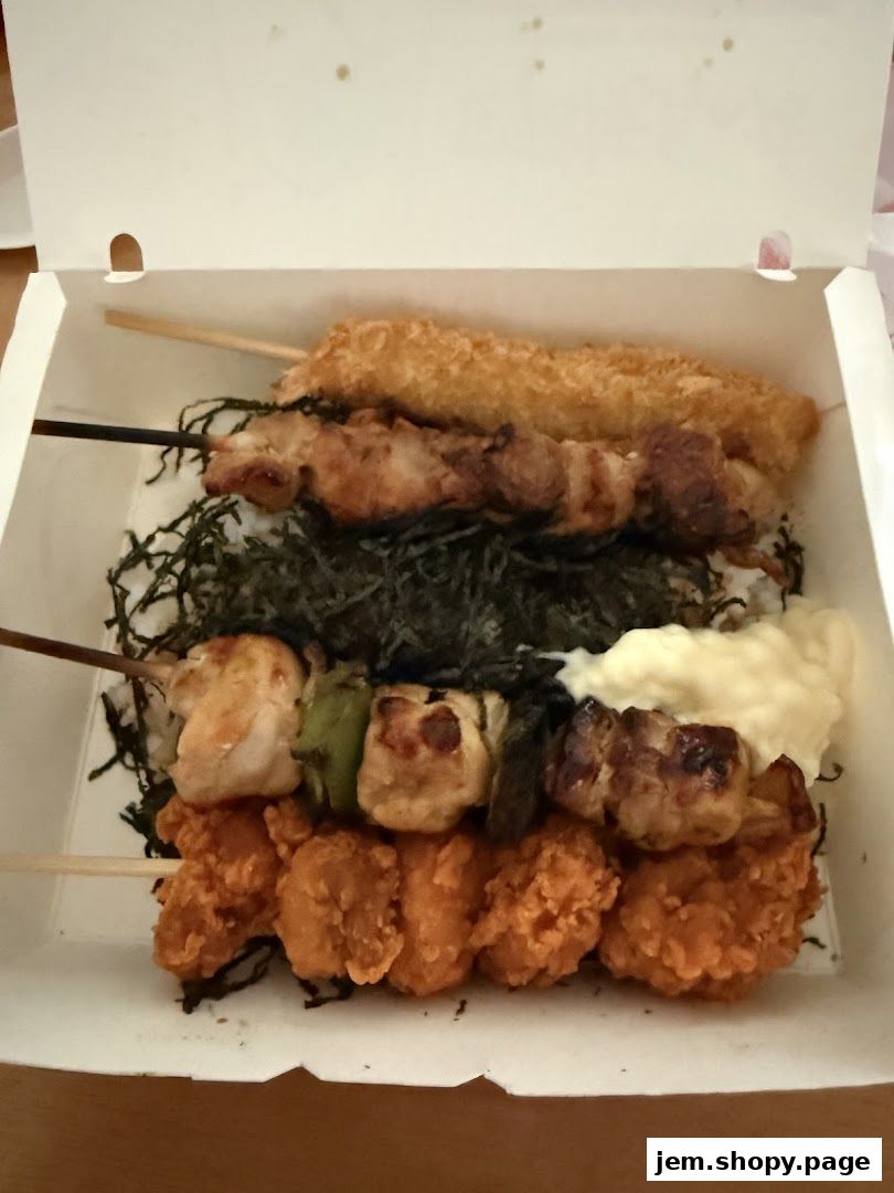 A takeaway box filled with assorted grilled skewers and fried chicken pieces on rice.