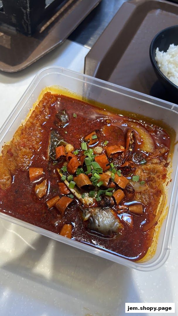 A takeout container filled with spicy fish stew, garnished with chili peppers and green onions.