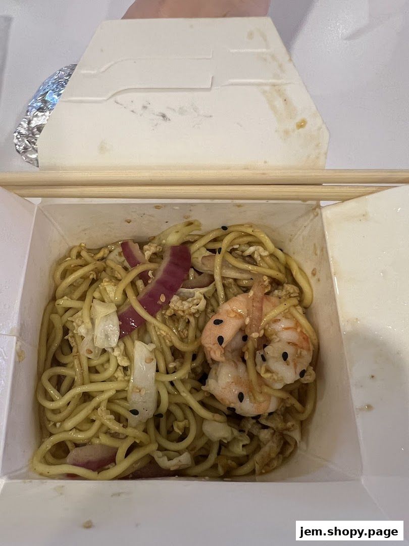 A close-up shot of a takeaway box filled with noodles, shrimp, and vegetables.