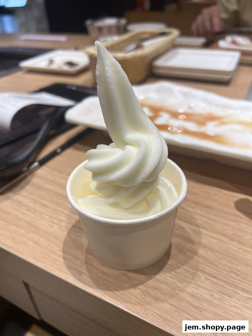 A cup of soft-serve ice cream sits on a wooden table, with plates in the background.