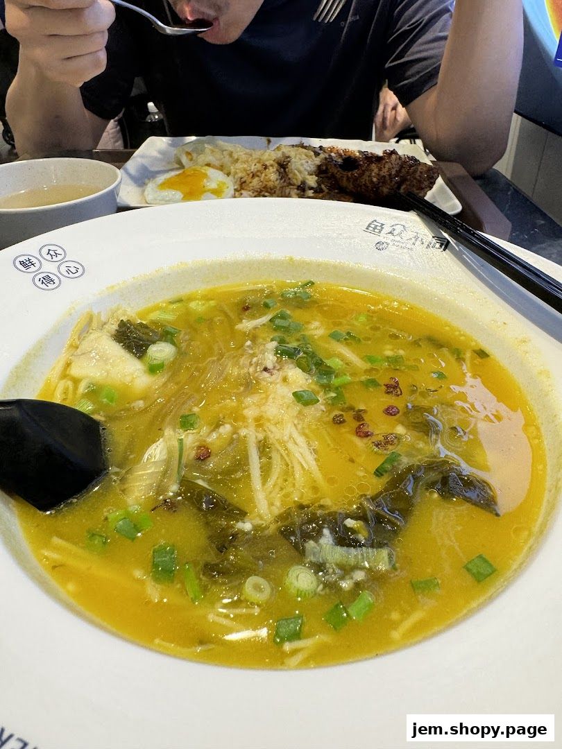 A person is eating a bowl of fish soup with noodles and a side of rice and fried egg.