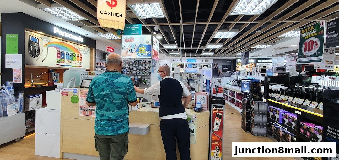Two people at a cashier counter in an electronics store with various brands displayed.