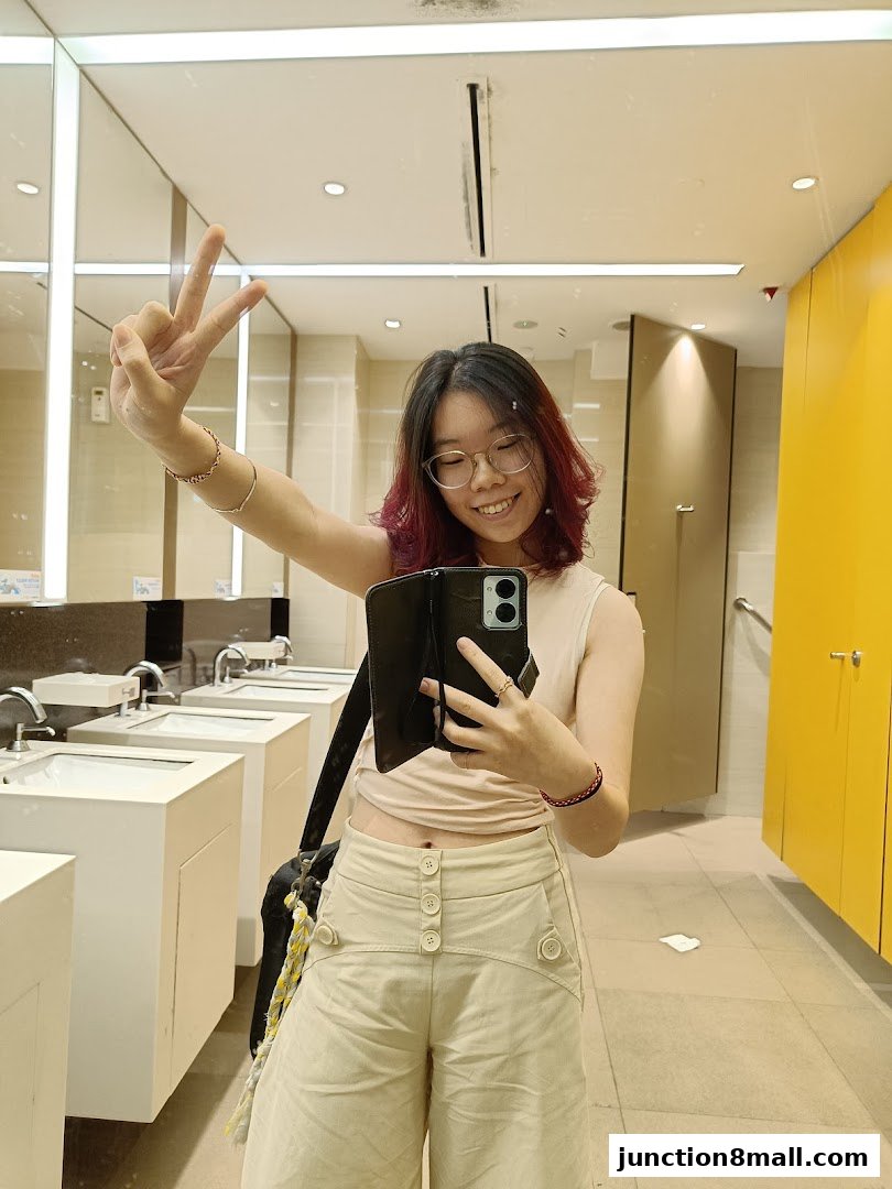 A young woman takes a mirror selfie in a clean, modern restroom, showing off her outfit.