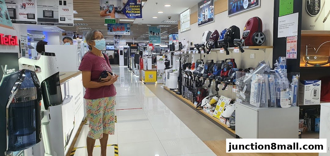 A woman stands in an electronics store aisle, surrounded by various appliances and displays.