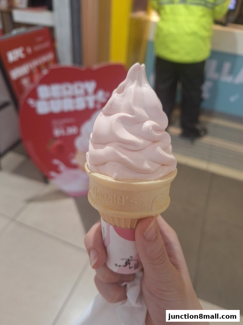 A hand holds a pink soft-serve ice cream cone with a promotional sign in the background.