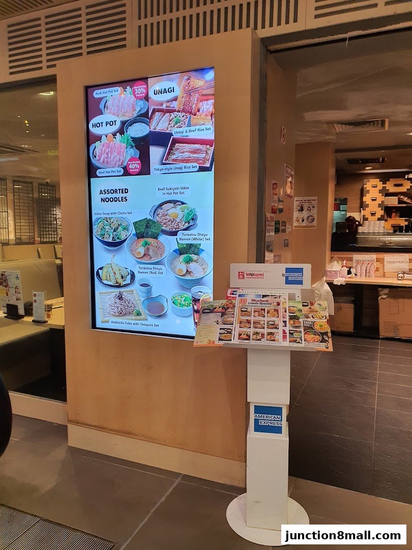 A digital menu board and a physical menu stand displaying Japanese food items at Watami.