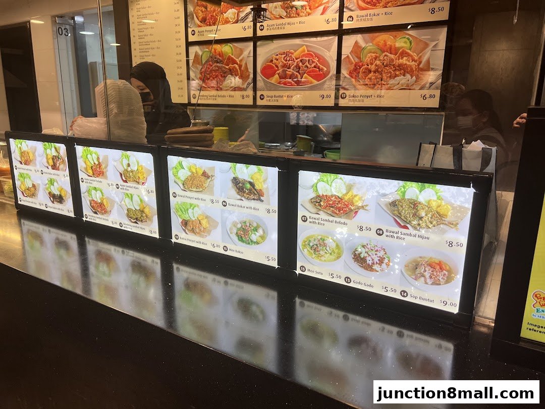 A food stall counter displaying illuminated menus with pictures of various dishes and prices.