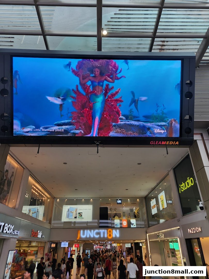 A large screen displays a movie trailer for 'The Little Mermaid' above a busy shopping mall walkway.