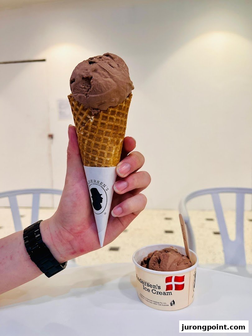 A hand holds a chocolate ice cream cone, with another cup of ice cream on a white table.
