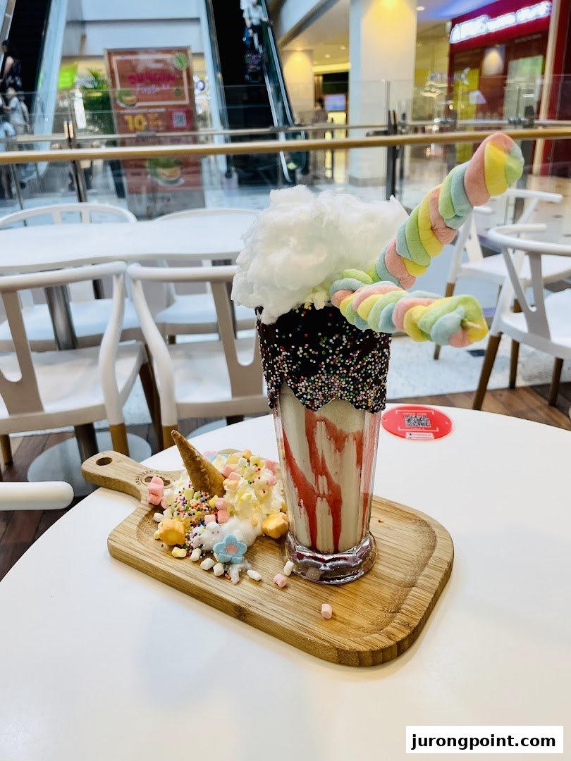 An elaborate milkshake topped with cotton candy, colorful marshmallows, and a unicorn-themed dessert on a wooden board.