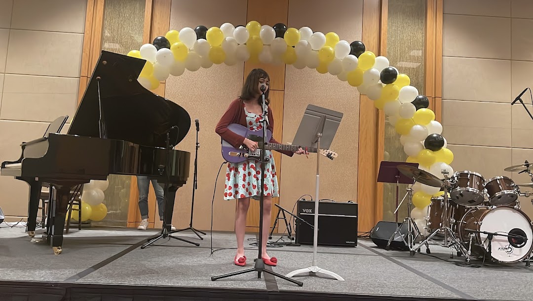 A young person performs live on stage with a guitar, accompanied by a piano and drums.