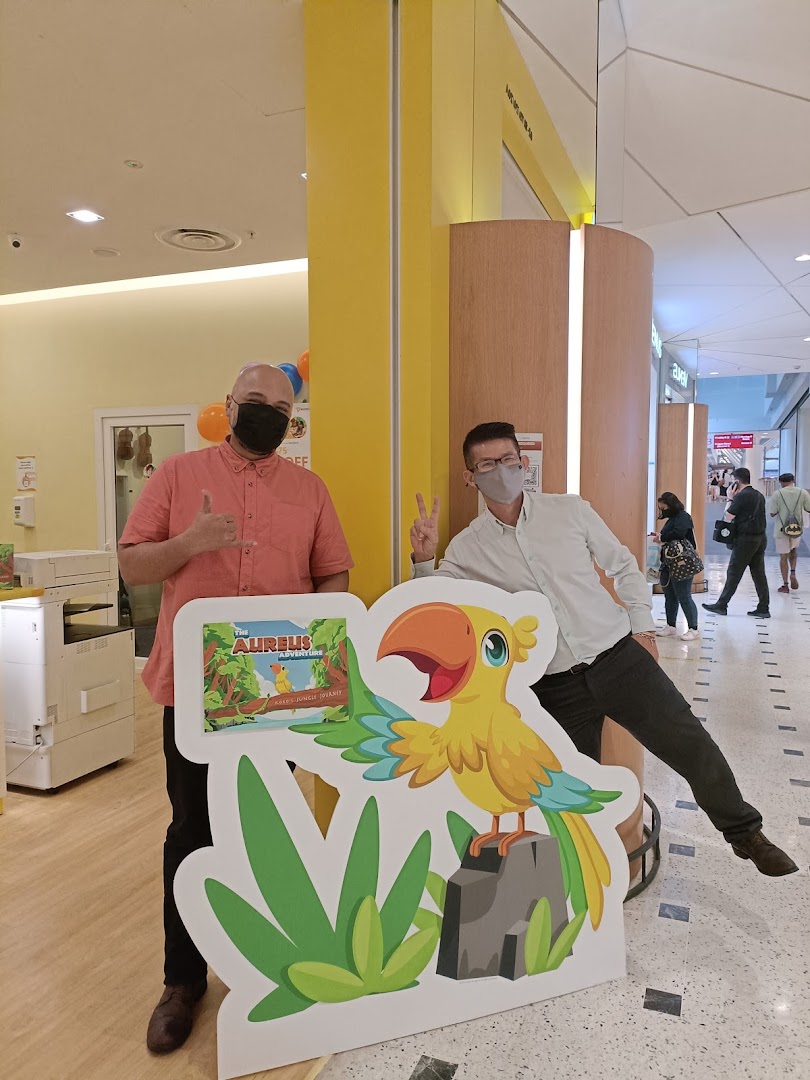 Two masked men pose with a colorful 'Aureus Adventure' standee inside a shopping mall.