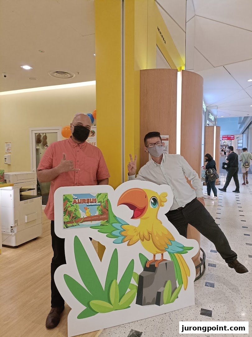 Two masked men pose with a colorful 'Aureus Adventure' standee inside a shopping mall.