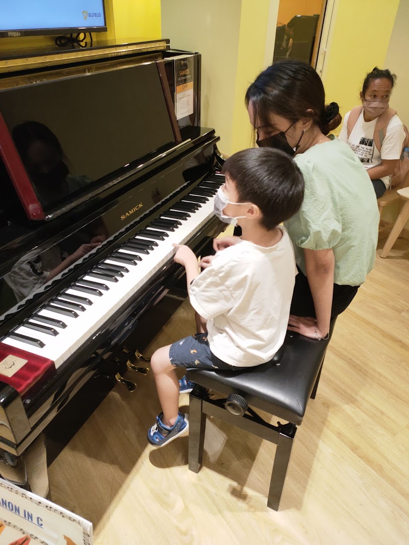 A child practices piano with an adult's guidance at Aureus Academy, with another person in the background.
