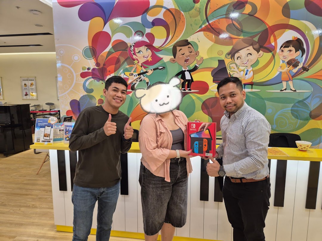 Three people, one receiving a Nintendo Switch, in a vibrant music academy setting with a piano-themed counter.