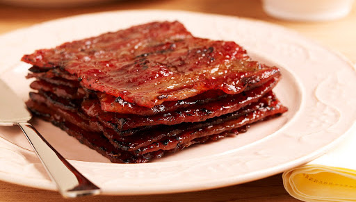 A stack of glossy, caramelized bak kwa (barbecued meat slices) on a light pink plate with a serving spatula.